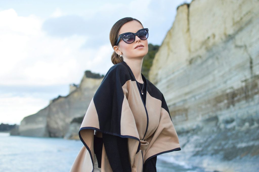 Femme portant une robe noire et marron avec des lunettes de soleil, debout près d'un plan d'eau sous un ciel blanc et bleu.